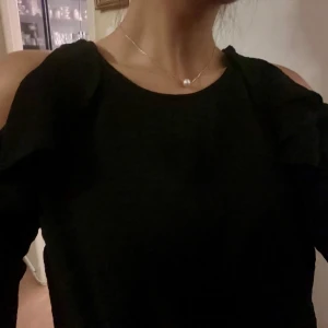 Svart blus med volangdetaljer - Snygg svart blus med volangdetaljer på ärmarna. Blusen har en offshoulder-design som ger en trendig look. Perfekt för att styla upp din outfit med en elegant touch. Använd 1 gång! Så den är nästan som nytt skick! 