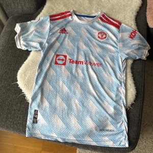 Manchester United fotbollströja - Säljer en Manchester United fotbollströja från Adidas med Ronaldo 7 på ryggen. Tröjan är vit med blått mönster och har röda detaljer. Den är kortärmad och har klubbens emblem på bröstet samt TeamViewer som sponsorlogga. Storlek 176