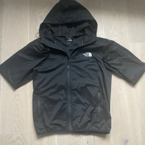 Svart vindjacka från The North Face - Säljer en svart vindjacka från The North Face med korta ärmar och huva. Jackan har en dragkedja framtill och är perfekt för blåsiga dagar. Den är tillverkad i ett lätt syntetmaterial och har en stilren design med logotyp på bröstet.
