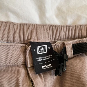 Beige byxor från Sweet Skibs - Snygga beige byxor från Sweet Skibs med en praktisk svart bälte. Byxorna har en avslappnad passform och är perfekta för en casual look. De har en dragkedja och fickor både fram och bak.