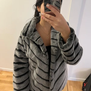 Grå pälsjacka med svarta ränder - Snygg och trendig grå pälsjacka med svarta ränder. Jackan har en fluffig och mjuk textur som ger en lyxig känsla. Perfekt för att hålla sig varm och stilren under kallare dagar.