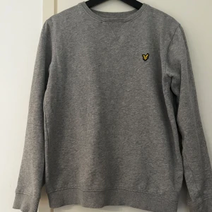 Grå sweatshirt från Lyle & Scott - Säljer en stilren grå sweatshirt från Lyle & Scott med deras ikoniska logga på bröstet. Tröjan har långa ärmar och ribbade muddar vid ärmslut och nederkant. Perfekt för en avslappnad look.