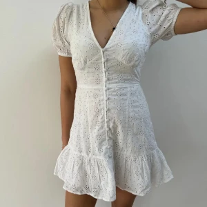 Vit klänning  - Vit klänning från Gina Tricot med vackert broderi. Den har korta puffärmar och en volangdetalj vid fållen. Klänningen är v-ringad och har knappar framtill. Perfekt för en somrig look! 🌸helt oanvänd 
