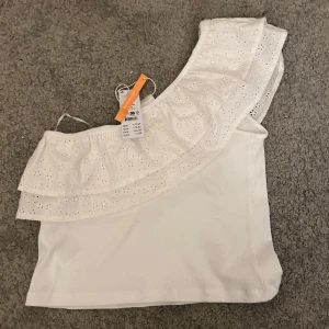 Vit oneshoulder topp med volang - Säljer en söt vit oneshoulder topp från Gina Tricot med volangdetaljer. Toppen har ett broderat mönster och är perfekt för varma dagar. Den är i ett ribbat material som ger en skön passform. Den är helt ny och nypris är 179 kr💕passar mig som har S!!