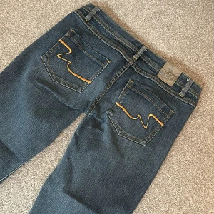 Mörkblå jeans med broderad detalj - Snygga mörkblå jeans med broderad detalj på insidan av linningen och kontrastsömmar på bakfickorna. Perfekta för en avslappnad stil med en touch av unik design.