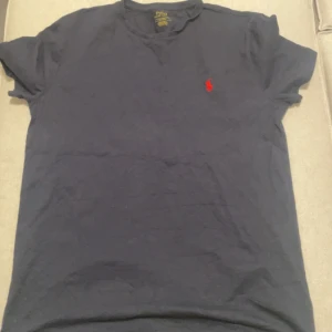 Ralph Lauren t-shirt  - Säljer en klassisk mörkblå t-shirt från Polo Ralph Lauren med det ikoniska röda broderade logotypen på bröstet. T-shirten har en normal passform och är tillverkad i mjuk bomull. Perfekt för en stilren och avslappnad look.