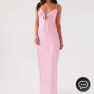 Rosa långklänning med knyt - Säljer en elegant rosa långklänning med smala axelband som är justerbara och knytning fram och bak. Klänningen har en öppen rygg och är tillverkad i ett glansigt material som ger en lyxig känsla. Perfekt för balen, bröllop eller midsommar!