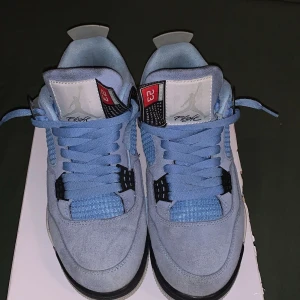 Jordan 4  - Jordans i ljusblå färg med svarta och grå detaljer. Skorna har snörning och en ikonisk Jumpman-logga på hälen. Perfekta för den som vill ha en stilren och sportig look.