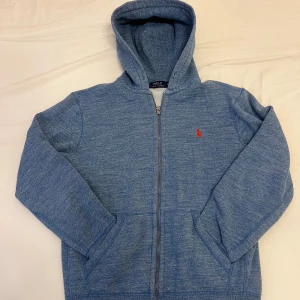 Blå zip up hoodie från Polo Ralph Lauren - Säljer en snygg blå hoodie från Polo Ralph Lauren med dragkedja och huva. Den har en röd logotyp på bröstet och är tillverkad i ett mjukt material. Perfekt för en avslappnad stil.