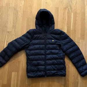 Lyle & Scott dunjacka - Säljer en svart pufferjacka från Lyle & Scott med huva och dragkedja. Hade för mig att den skulle läcka lite ludd från något ställe, men efter mycket sökande fann jag inget. 