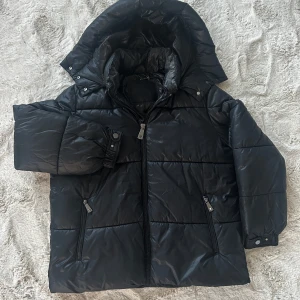 Svart dunjacka från Guess - Säljer en svart dunjacka från Guess med huva och dragkedja. Jackan har en quiltad design och praktiska fickor med dragkedjor. Perfekt för kyliga dagar.