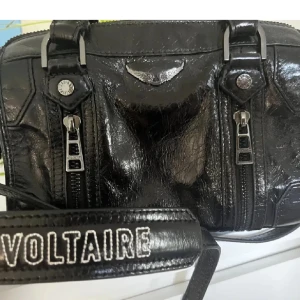 Svart handväska från Zadig & Voltaire - Snygg svart handväska i skinn från Zadig & Voltaire med dubbla dragkedjor framtill och justerbar axelrem. Storlek xs men rymmer mycket! Perfekt för att ge din outfit en edgy touch. kan tänka mig gå ner i pris! 