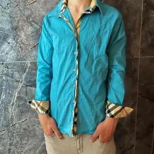 Turkos Burberry skjorta - Snygg turkos skjorta med långa ärmar och unika rutiga detaljer på insidan av kragen och ärmsluten. Perfekt för att ge en färgklick till din outfit. Knappar framtill för enkel stängning.