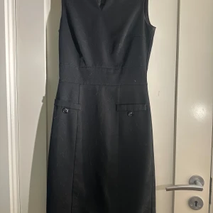 Svart ärmlös klänning - Elegant svart ärmlös klänning med v-ringning och dekorativa knappar framtill. Perfekt för en stilren look.