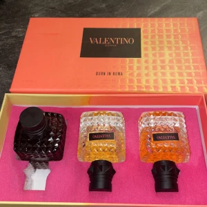 Valentino Donna Born in Roma parfymset - Exklusivt parfymset från Valentino med tre flaskor i elegant design. Två flaskor har en klar glasdesign med gyllene och orange toner, medan den tredje är mörkare. Förpackningen är lyxigt rosa med svart och guld detaljer.                                   OBS: den svarta har läckt, finns ungefär 1/4 kvar av den