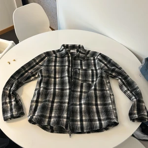 Svartvit rutig overshirt - Säljer en stilren svartvit rutig overshirt med dragkedja. Perfekt för lager-på-lager under höst och vår. Den har en bröstficka och långa ärmar. Passar både till vardags och mer avslappnade tillfällen.