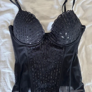 Svart paljettkorsett från H&M - Glittrig mesh korsett topp / festtopp, ger jättefin form. Köpt secondhand men passade inte till det tillfället jag tänkt! Skickar jättegärna bild på, bara att höra av sig 🙏😇 Från H&M i storlek 40 men skulle säga att den är mer 38/36 beroende på passform. Glittrande paljetter framtill. Den har justerbara axelband och en hakknäppning bak. Perfekt för en kväll ute eller en speciell tillställning. ✨