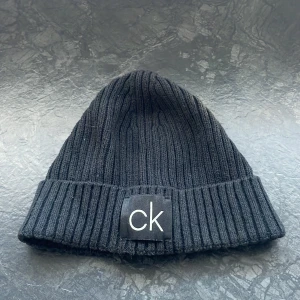 Svart ribbad mössa från Calvin Klein - Snygg svart ribbad mössa från Calvin Klein med ett stilrent 'ck' märke framtill. Perfekt för att hålla värmen under kyliga dagar. Nypris 350kr Mössan är tillverkad i ett mjukt och bekvämt material ✅