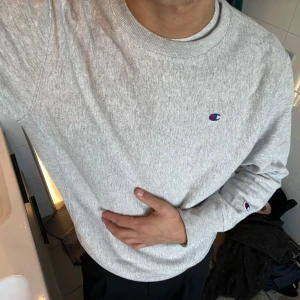 Grå sweatshirt från Champion - Säljer en klassisk grå sweatshirt från Champion med deras ikoniska logga på bröstet och ärmen. Tröjan har långa ärmar och en rund halsringning. Perfekt för en avslappnad stil.
