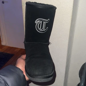 Svarta boots med broderad logotyp - Snygga svarta boots i mocka med en broderad logotyp på skaftet. Perfekta för att hålla fötterna varma under kyliga dagar. De har en klassisk design med rund tå och är fodrade för extra komfort.