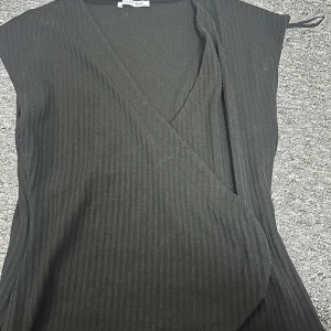 Svart omlotttopp från Zara - Snygg svart omlottklänning från Zara Trafaluc med ribbad textur. Klänningen är ärmlös och har en stilren design som passar perfekt för olika tillfällen. Tillverkad i Turkiet.