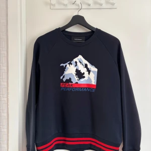 Mörkblå tröja från Peak Performance - Säljer en mörkblå tröja från Peak Performance med ett bergsmotiv och texten 'Peak Performance' i rött och blått. Tröjan har långa ärmar och röd-svart randig detalj vid nederkanten. Perfekt för en sportig look!