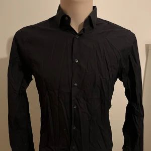 Svart skjorta från The Shirt Factory - Snygg svart skjorta från The Shirt Factory med slim fit passform. Skjortan har långa ärmar och knappar framtill. Perfekt för en stilren look.