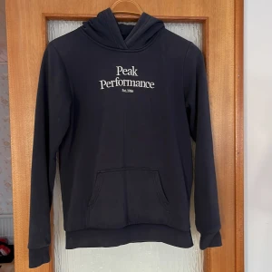 Marinblå hoodie från Peak Performance - Säljer en stilren marinblå hoodie från Peak Performance med vit logga på bröstet. Tröjan har en klassisk känguruficka och långa ärmar. Perfekt för en avslappnad look.