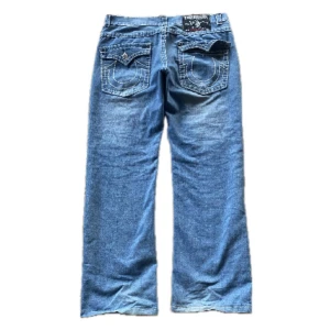 Blå jeans från True Religion - Säljer ett par klassiska blå jeans från True Religion med snygga broderade detaljer på bakfickorna. Jeansen har en rak passform och är tillverkade i USA. Perfekta för en avslappnad stil. En av knapparna på bak fickan är av.