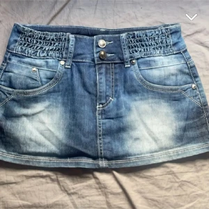 Blå jeanskjol från Toxik3 - Snygg blå jeanskjol från Toxik3 med smockdetaljer i midjan och dubbla knappar fram. Kjolen har en kort längd och klassiska femficksdetaljer. Perfekt för en avslappnad stil.