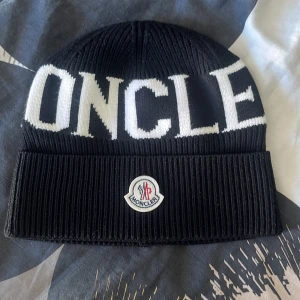 Svart mössa från Moncler - Snygg svart mössa från Moncler med vit text och logotyp framtill. Den är ribbad och har en klassisk design som passar perfekt för kyligare dagar.