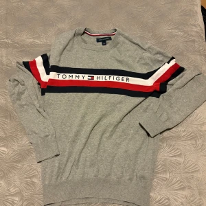 Grå tröja från Tommy Hilfiger - Snygg grå tröja från Tommy Hilfiger med deras ikoniska logga i marinblått, rött och vitt över bröstet. Tröjan har långa ärmar och ribbade muddar vid ärmslut och nederkant. Perfekt för en stilren look.