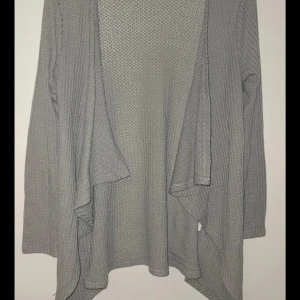 Grå stickad kofta - Säljer en mysig grå stickad kofta med långärmad design. Perfekt för kyliga dagar och enkel att matcha med olika outfits. Koftan har en lös passform och ett stilrent utseende.