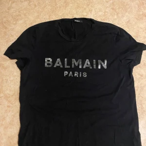 Svart t-shirt från Balmain - Snygg svart t-shirt från Balmain med deras ikoniska logga i silver på bröstet. Perfekt för en stilren och modern look. Tillverkad i mjukt bomullsmaterial för extra komfort.