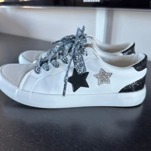 Vita sneakers med stjärndetaljer - Snygga vita sneakers med glittriga svarta och silverstjärnor på sidan. Skorna har leopardmönstrade snören och glittriga svarta detaljer på hälen. Perfekta för att ge din outfit en extra touch.
