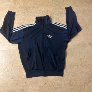 Mörkblå träningsjacka från Adidas - Säljer en snygg mörkblå träningsjacka från Adidas med de klassiska tre ränderna längs ärmarna. Jackan har dragkedja framtill och två fickor. Perfekt för en sportig look! 💙