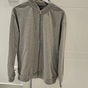 Grå tröja med dragkedja från DeFacto - Säljer en stilren grå tröja från DeFacto med dragkedja framtill. Tröjan har en enkel design med långa ärmar och ribbade muddar vid ärmslut och nederkant. Perfekt för en avslappnad look.