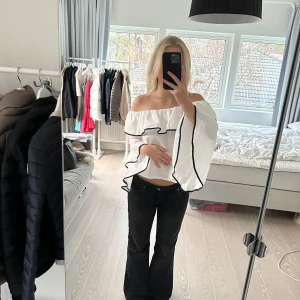 Vit offshoulder blus från Zara - Säljer en snygg vit offshoulder blus från Zara med svarta detaljer. Blusen har volanger och vida ärmar som ger en trendig look. Perfekt för att styla med jeans eller kjol. Hittar inte storleken men uppskattar till en S. Bra skick!