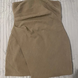 Beige axelbandslös klänning från Boohoo - Snygg beige axelbandslös klänning från Boohoo med en stilren design. Klänningen har en dragkedja baktill för enkel påklädning och en elegant omlott-detalj framtill. Perfekt för en chic look!