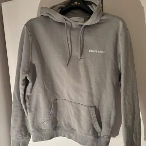 hoodie från Axel Arigato - Säljer en stilren grå hoodie från Axel Arigato med tryck 'ARIGATO ©2014' på bröstet och ryggen. Tröjan har en klassisk känguruficka och justerbar huva med dragsko. Perfekt för en avslappnad look.