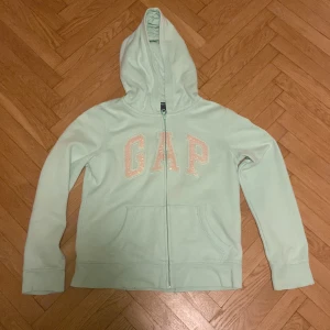 Ljusgrön hoodie från GAP - Säljer en ljusgrön hoodie från GAP med dragkedja och stor logga framtill. Tröjan har långa ärmar och en praktisk huva. Perfekt för en avslappnad stil. Står väldigt konstigt i storlekarna då den är köpt i Usa men skulle uppskatta är den är i storlek s här i Sverige.