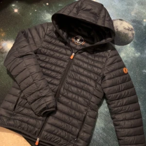 Svart pufferjacka från Save the Duck - Säljer en svart pufferjacka från Save the Duck i storlek S. Jackan är tillverkad med Plumtech syntetiskt dun och är 100% djurfri. Den har en huva och dragkedja framtill. Perfekt för kyliga dagar.