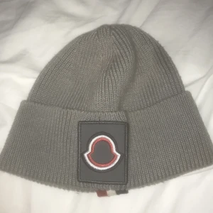 Grå stickad mössa från Moncler - Snygg grå stickad mössa från Moncler med ett stort broderat märke framtill. Mössan har en uppvikt kant och en randig detalj i svart, vit och röd på baksidan. Perfekt för att hålla sig varm och stilren under kyliga dagar. Säljer för 450 men pris kan diskuteras