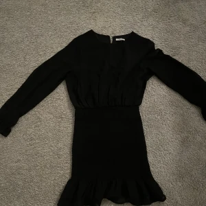 Svart klänning med volangdetalj - Elegant svart klänning med långa ärmar och volangdetalj vid fållen. Perfekt för en stilren look. Klänningen har en åtsittande passform i midjan och en rund halsringning.