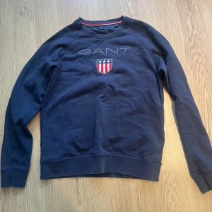 Mörkblå sweatshirt från GANT - Säljer en snygg mörkblå sweatshirt från GANT med ett broderat emblem på bröstet. Tröjan har långa ärmar och en rund halsringning med röd och vit randig detalj på insidan. Perfekt för en avslappnad stil. 176 cm i storlek men är stor som typ s.