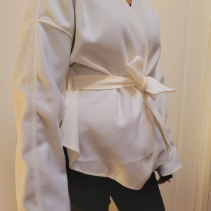 Vit omlottjacka med knytband - Säljer en stilren vit omlottjacka med knytband i midjan. Jackan har långa ärmar och en elegant v-ringad design. Perfekt för en chic och avslappnad look. Helt ny!