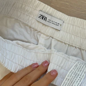 Vit kjol  från Zara - Snygg vit kjol från Zara med elastisk midja och snörning. Kjolen har praktiska dragkedjefickor fram och är perfekta för en avslappnad stil. Tillverkade i ett lätt och bekvämt material.