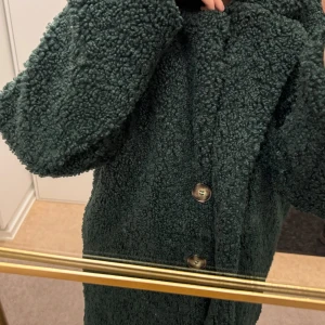 Grön teddyjacka med knappar - Mysig grön teddyjacka med knappar framtill. Perfekt för kyliga dagar med sin fluffiga och varma design. Jackan har långa ärmar och en klassisk krage.