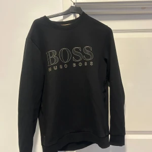 Svart sweatshirt från Hugo Boss Medium - Snygg svart sweatshirt från Hugo Boss med stort BOSS-tryck på framsidan. Tröjan har långa ärmar och en klassisk rund halsringning. Perfekt för en stilren och avslappnad look. I verkligheten är märket i mer guld!