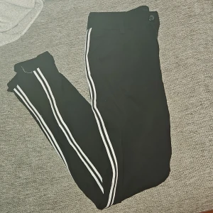 Svarta byxor med vita ränder - Snygga svarta byxor med vita ränder längs sidorna. Perfekta för en sportig look. Byxorna har en knapp i midjan och är bekväma att bära.